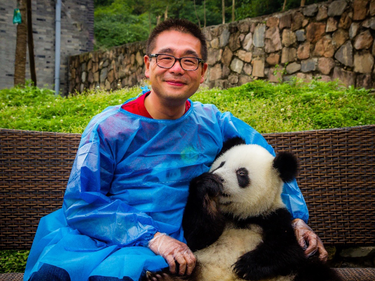 Leo Lo in a blue rain slicker smiling while holding a panda cub, sitting outdoors with a stone wall in the background.