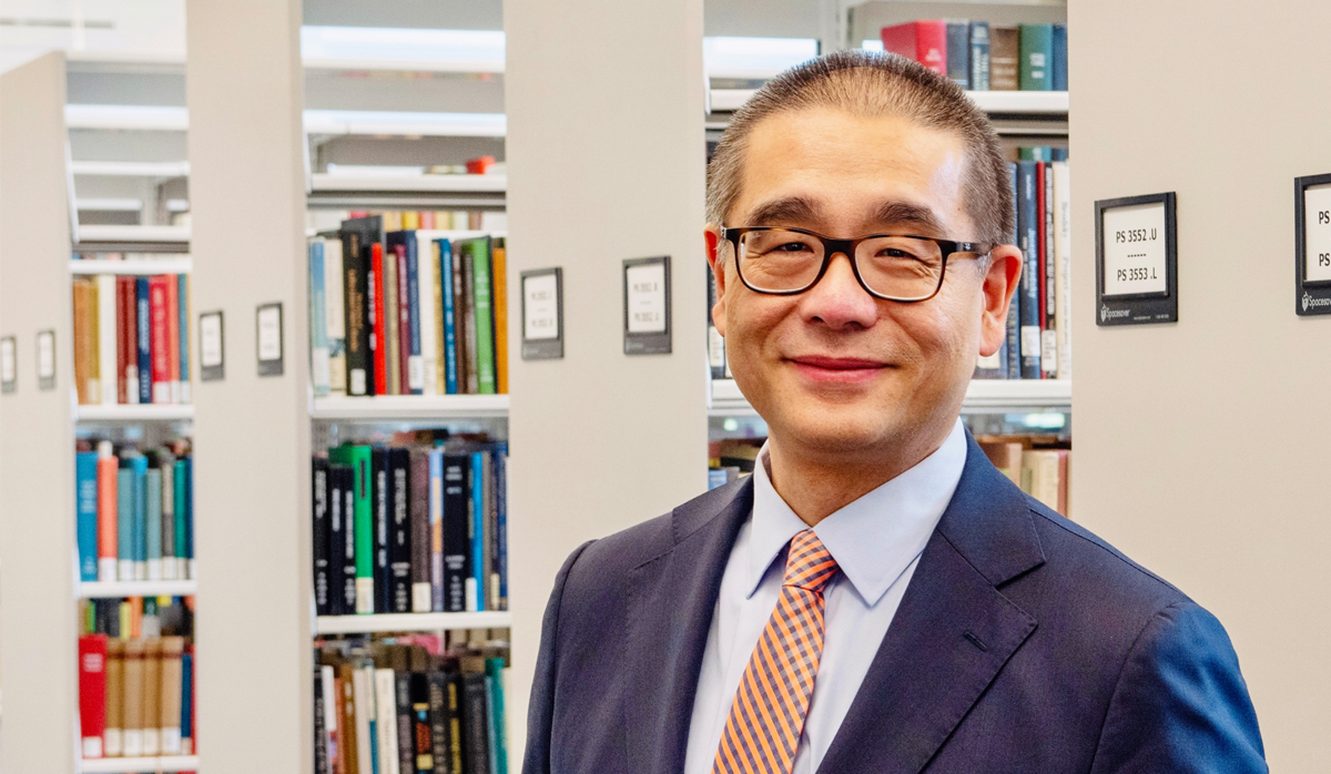 Dean Leo Lo of the UVA Library wearing a blue suit, orange and blue tie, and eyeglasses, standing in a library with shelves of books in the background.