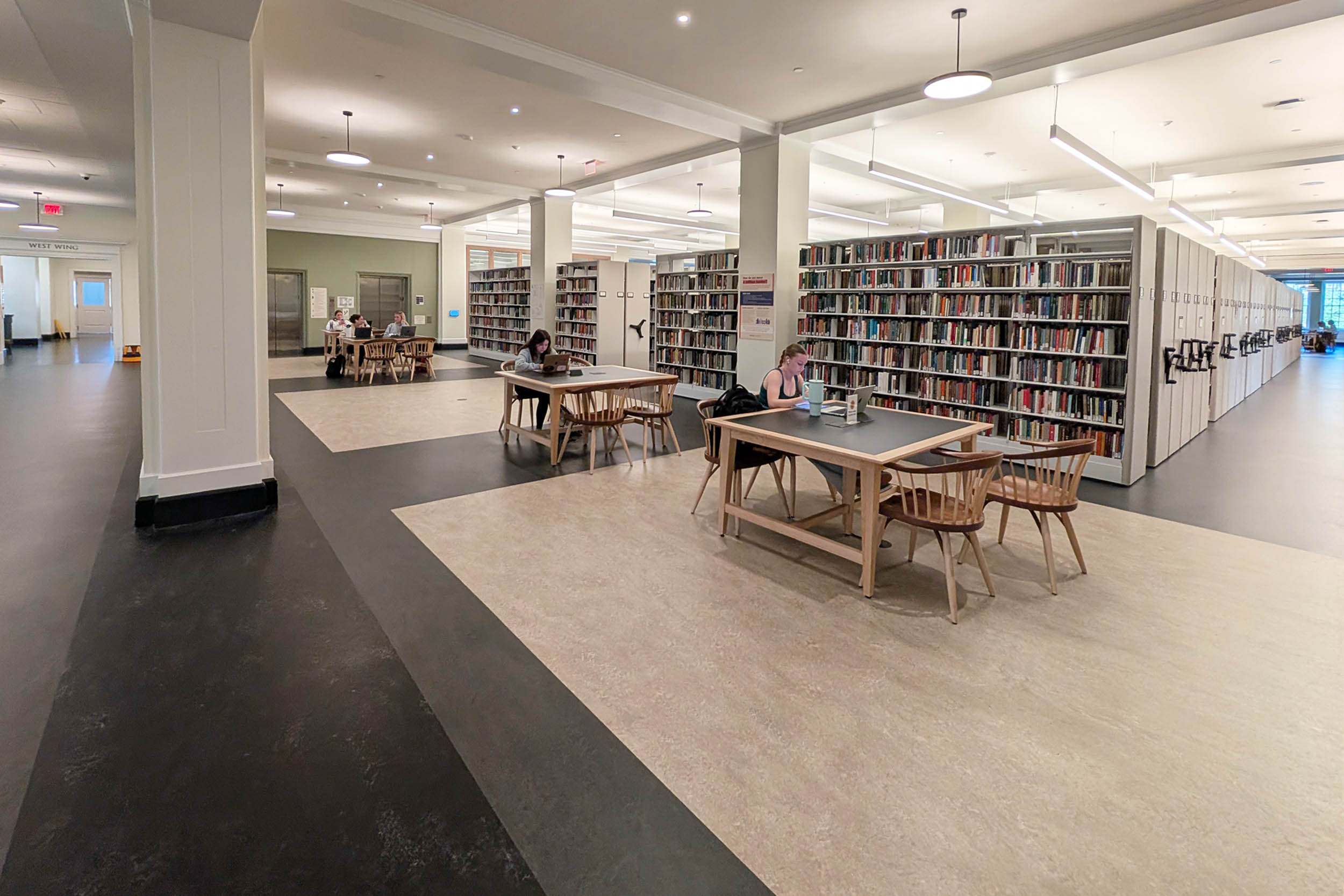 Interior of a library with patrons seated at tables reading and browsing books along rows of bookshelves. The space features ample lighting and a clean, organized layout.