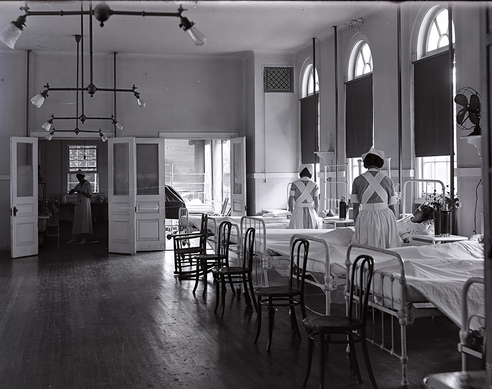 A dark room with a row of hospital beds and three nurses tending patients