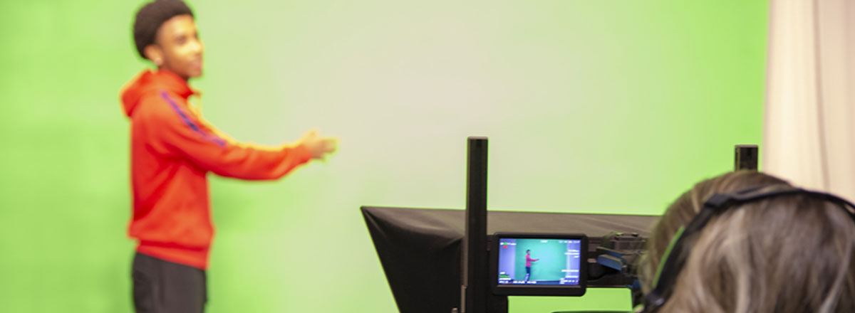 A student stands in front of a green screen while another operates a video camera