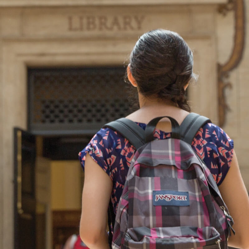 View of female student wearing backpack walking toward door marked Library