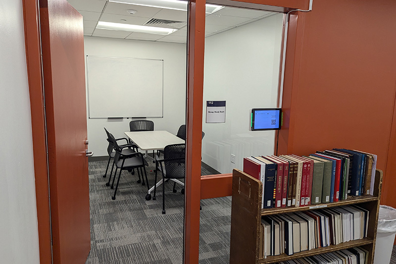 A small room has a digital screen and book cart outside the door. Inside the room is a table with seating for 6 and a white board on the wall. 