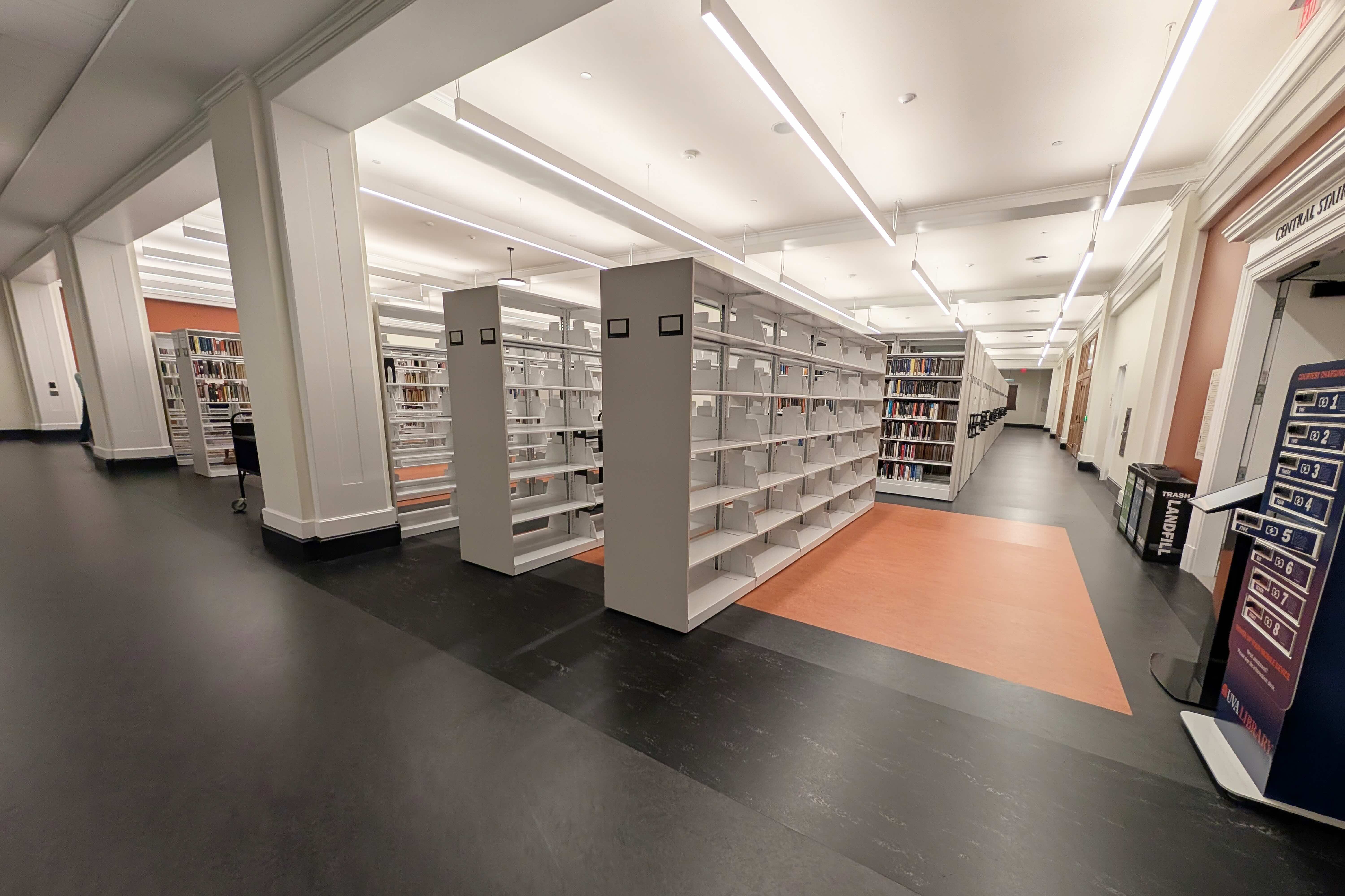 Interior of a library with rows of bookshelves filled with books, featuring a carpeted central walkway flanked by gray flooring. Bright overhead lighting illuminates the space.