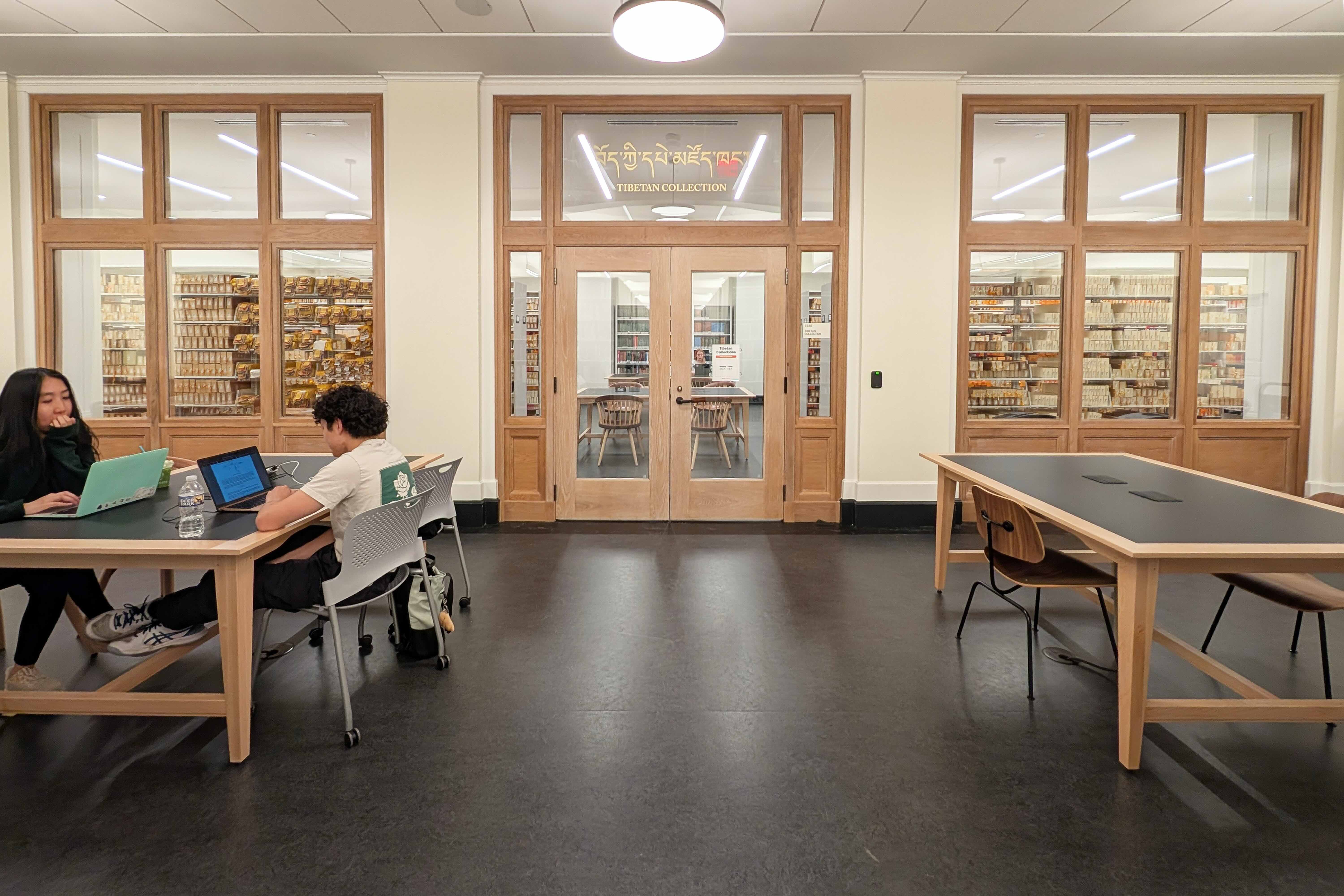 Two people seated at tables studying in a library hallway outside of a room. In the room shelves of books are visible through glass doors with gold text reading