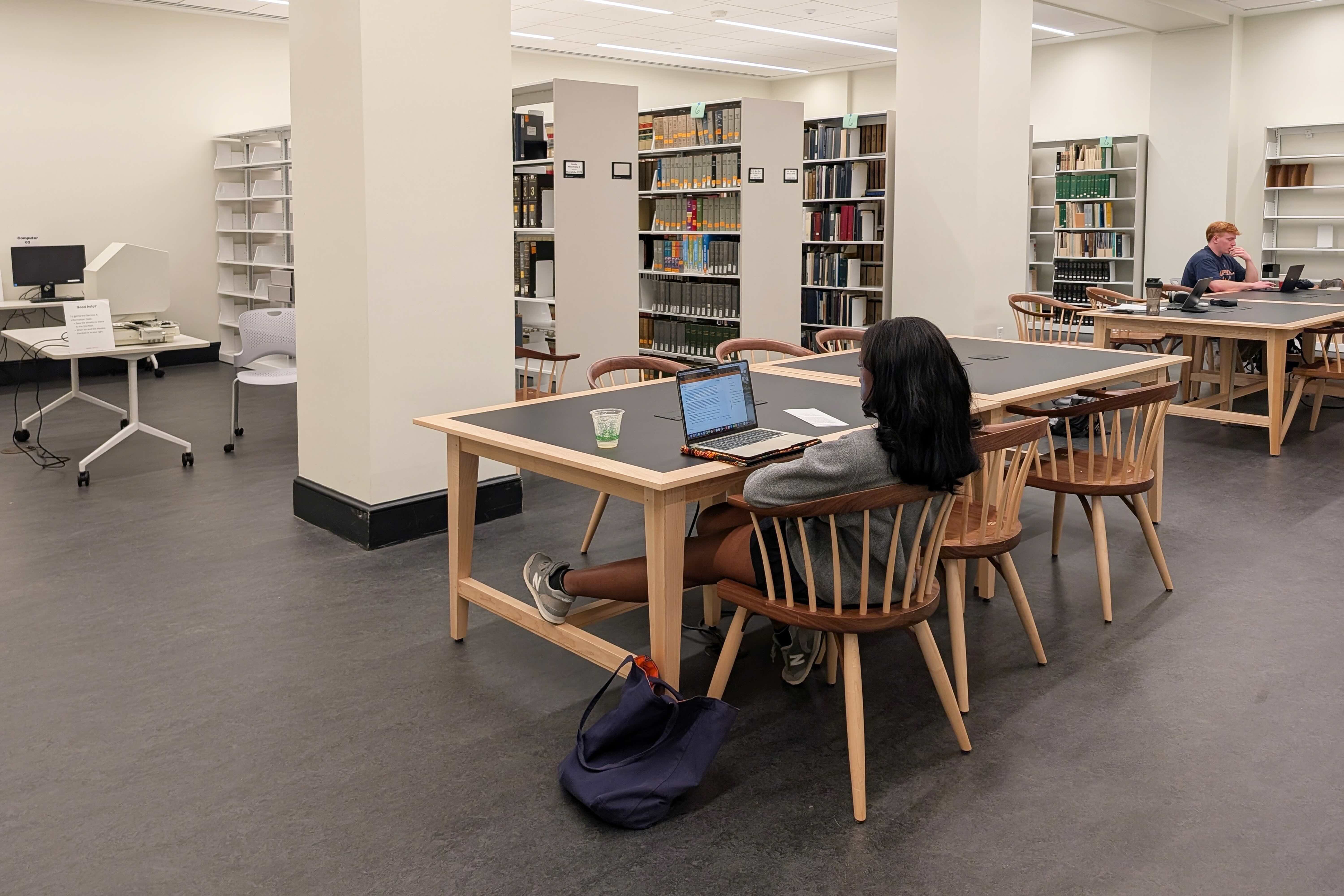 Person seated at a table working on a laptop in a modern library setting with bookshelves and other visitors in the background.
