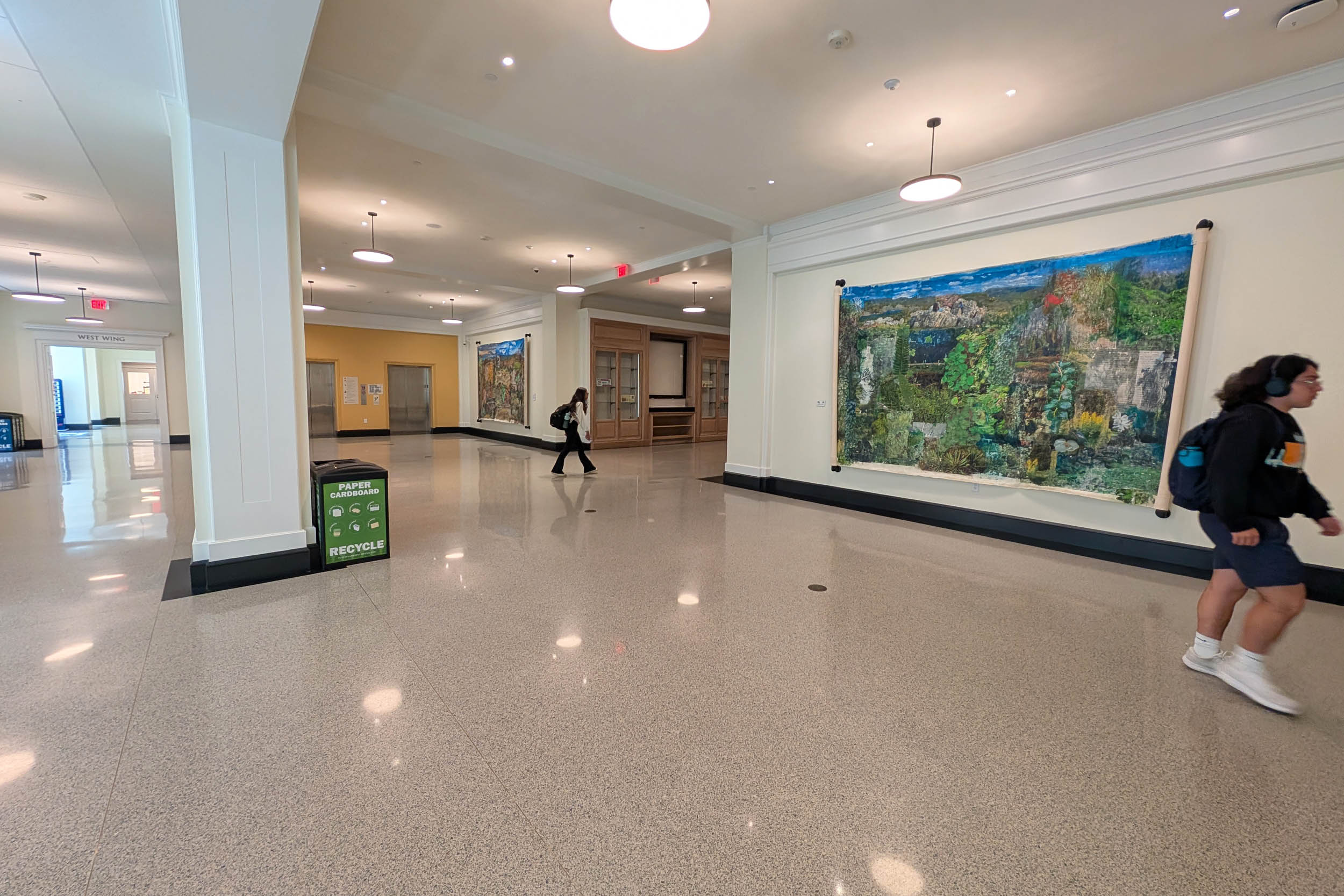 Interior of a spacious hallway with glossy tiled floors and large colorful art on the wall. People are moving through the area, and there are doors and signs visible.