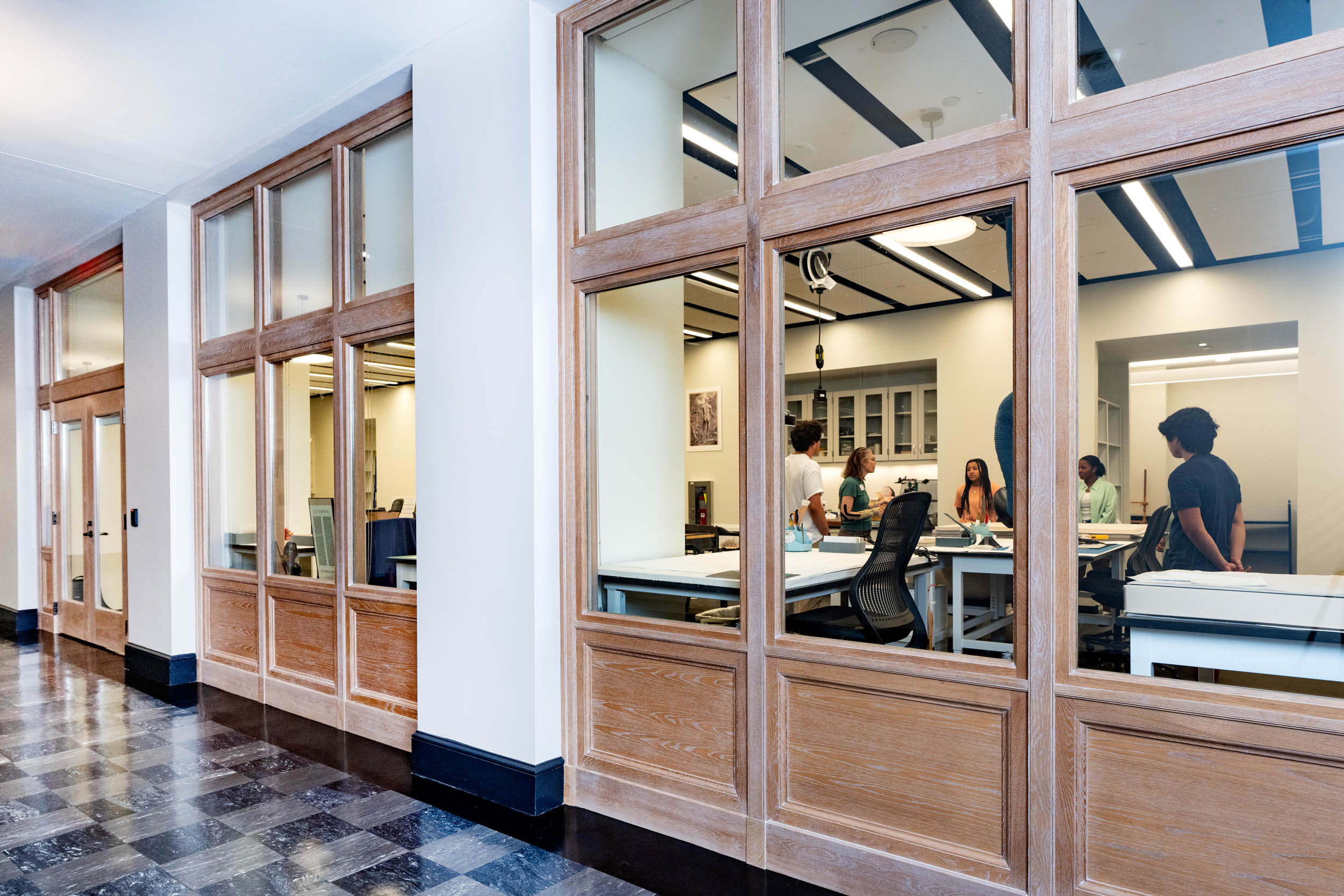 View of a large modern space seen through glass windows. The room has multiple desks and tables and a group of individuals are standing around a table having a discussion.