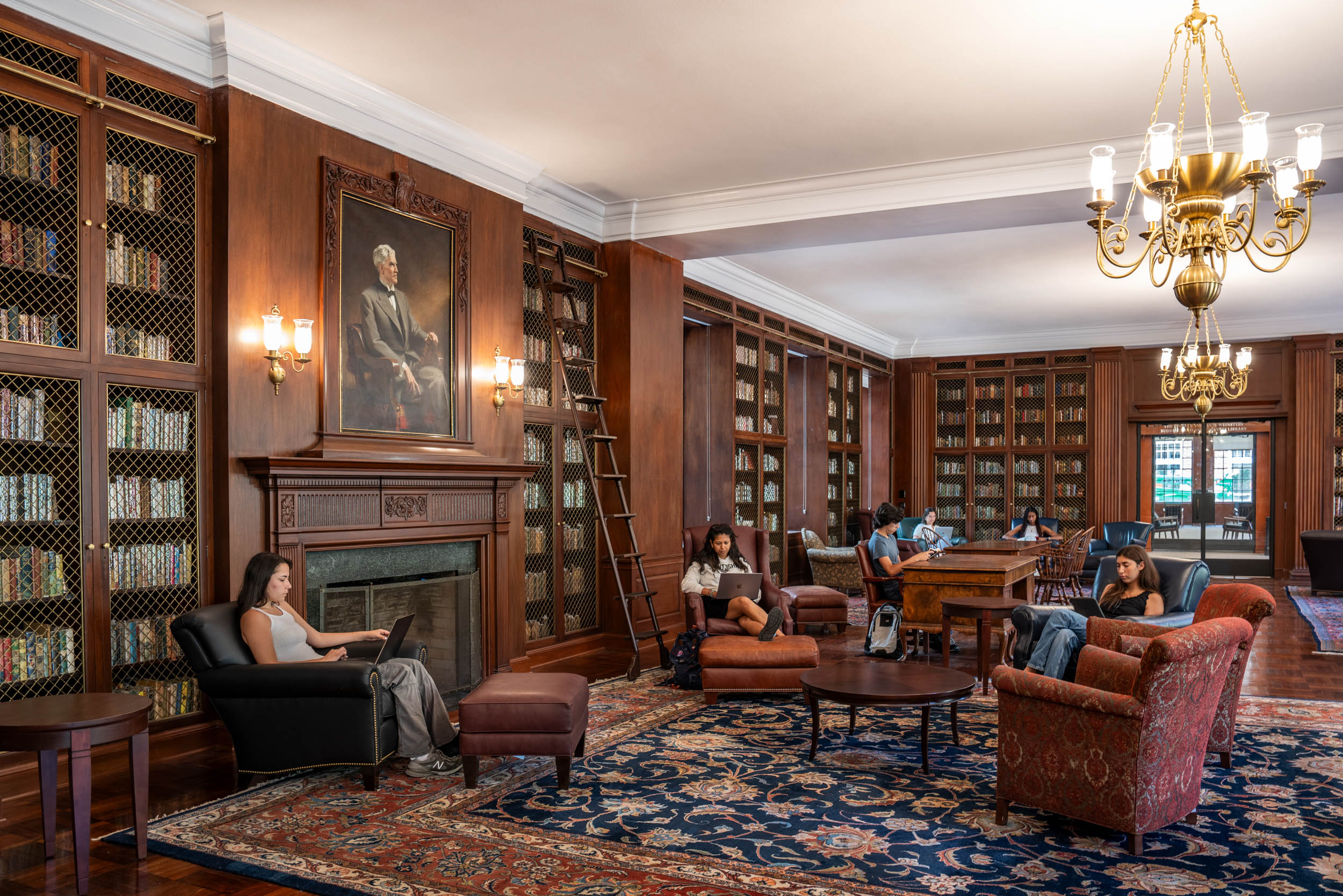 Interior of a traditional library with individuals reading and using laptops. The room features wood-paneled walls, floor-to-ceiling bookshelves, a large portrait above a fireplace, a chandelier, and ornate rugs.