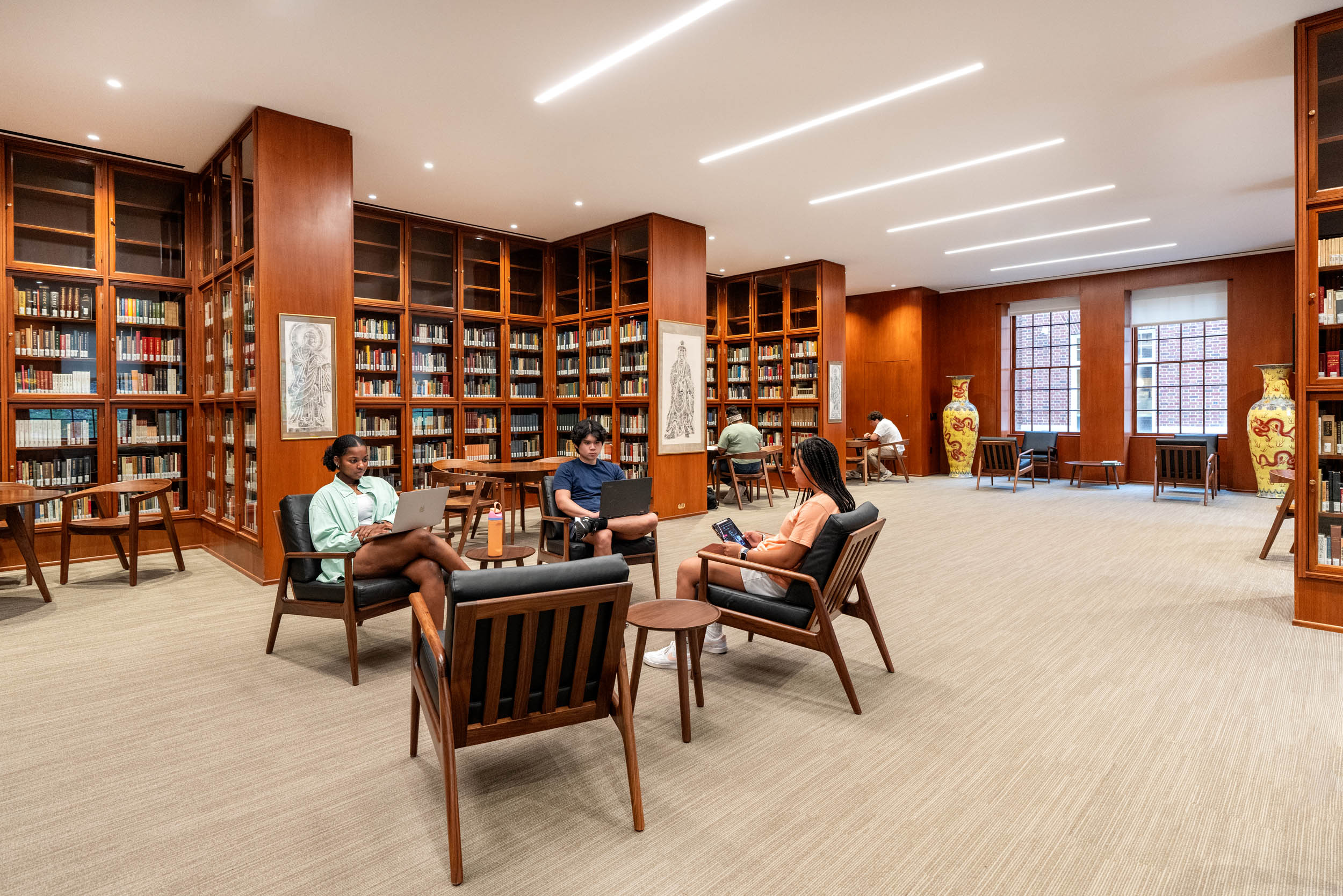 Three people sitting and reading in a well-lit library room with wood paneled walls and floor-to-ceiling bookshelves filled with books. There is comfortable seating and decorative items, including large vases and paintings.