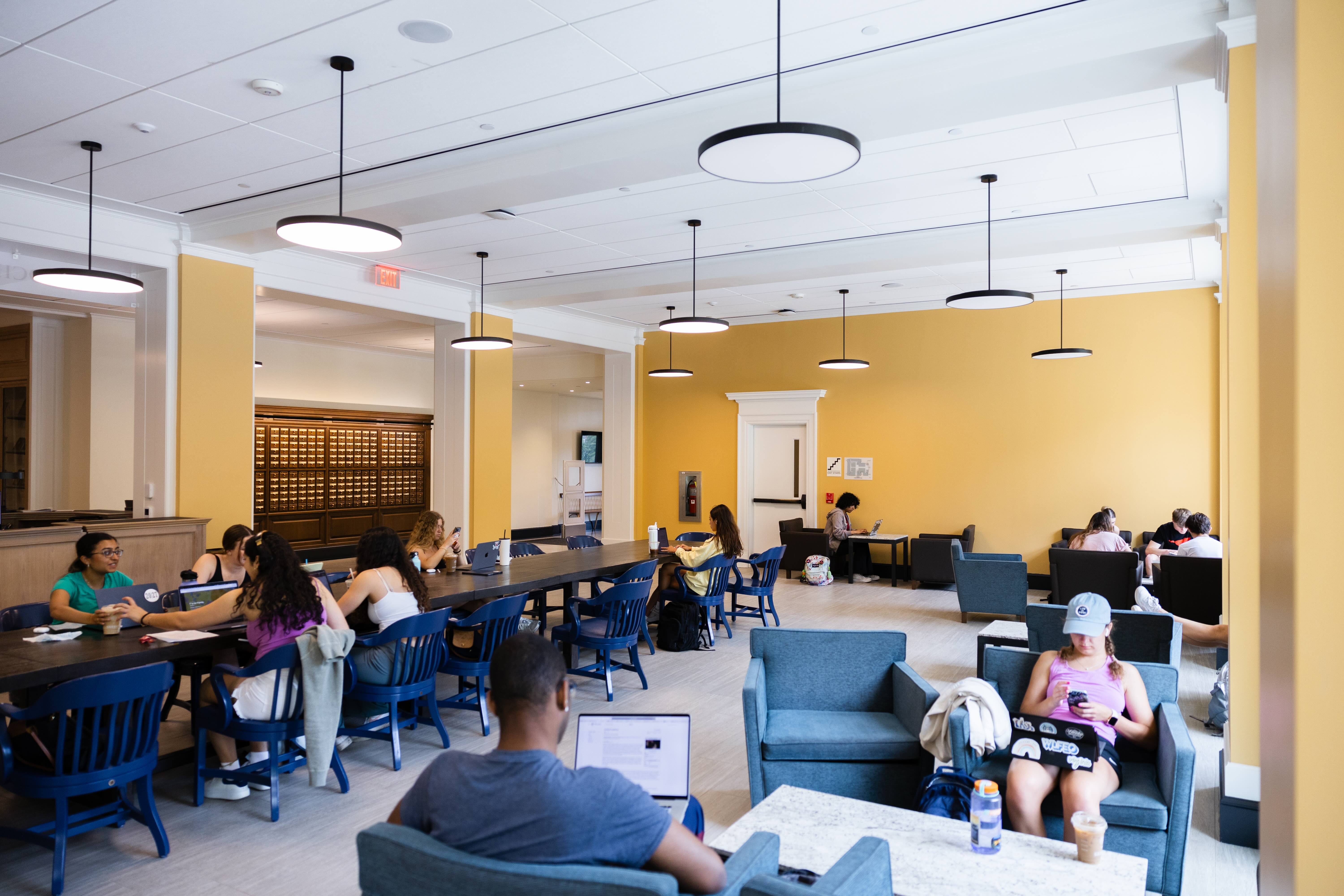 Students studying and using laptops in a brightly lit library reading room with yellow walls and modern furnishings.