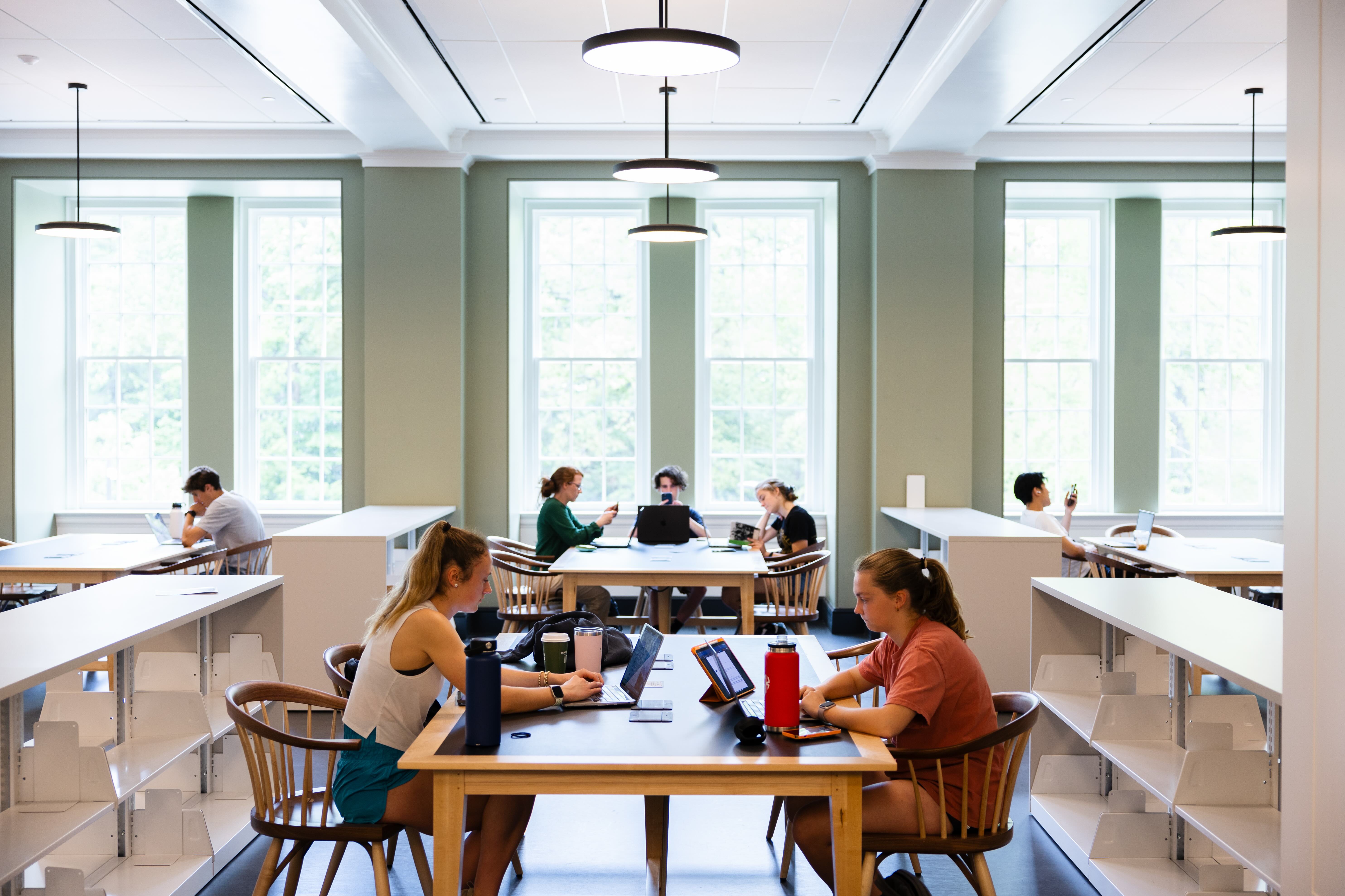 Students studying and working on laptops at tables in a bright, modern library with large windows and hanging circular lights.