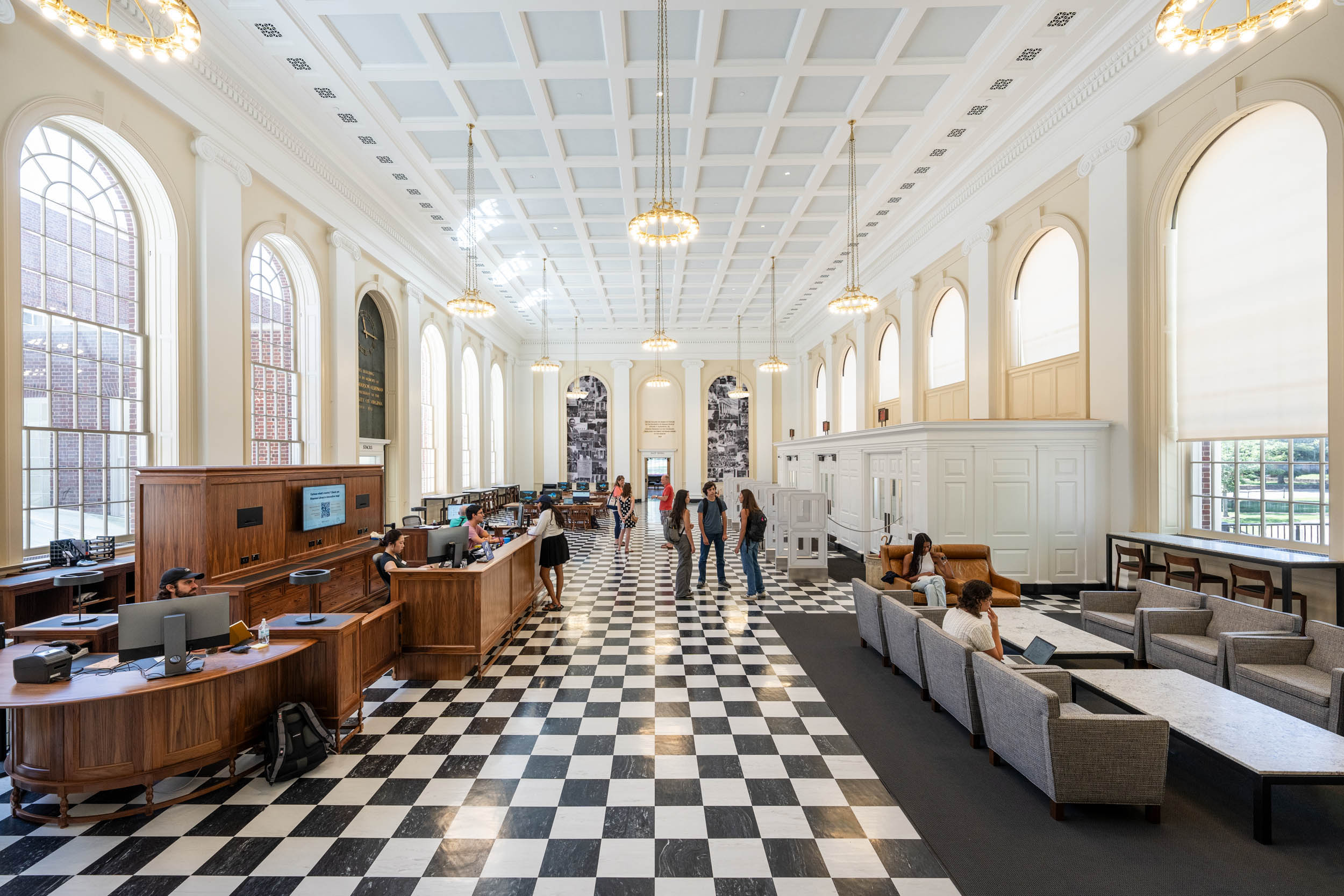 Interior of a spacious library with high ceilings, checkerboard flooring, large arched windows, and a variety of seating areas including desks and sofas. Several individuals are reading and working.