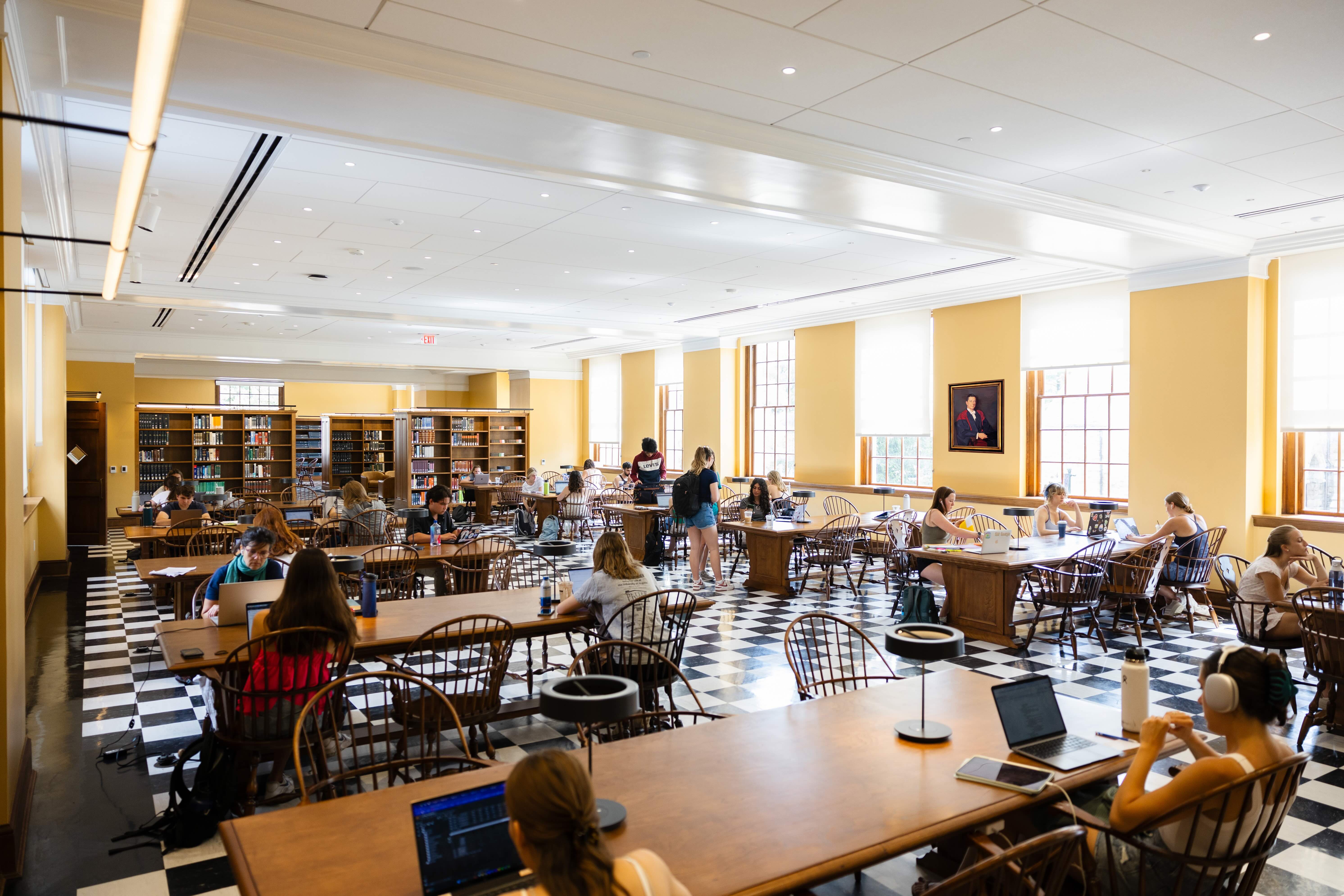 Interior of a busy library with numerous individuals studying and using laptops at tables, under bright lighting and surrounded by bookshelves.