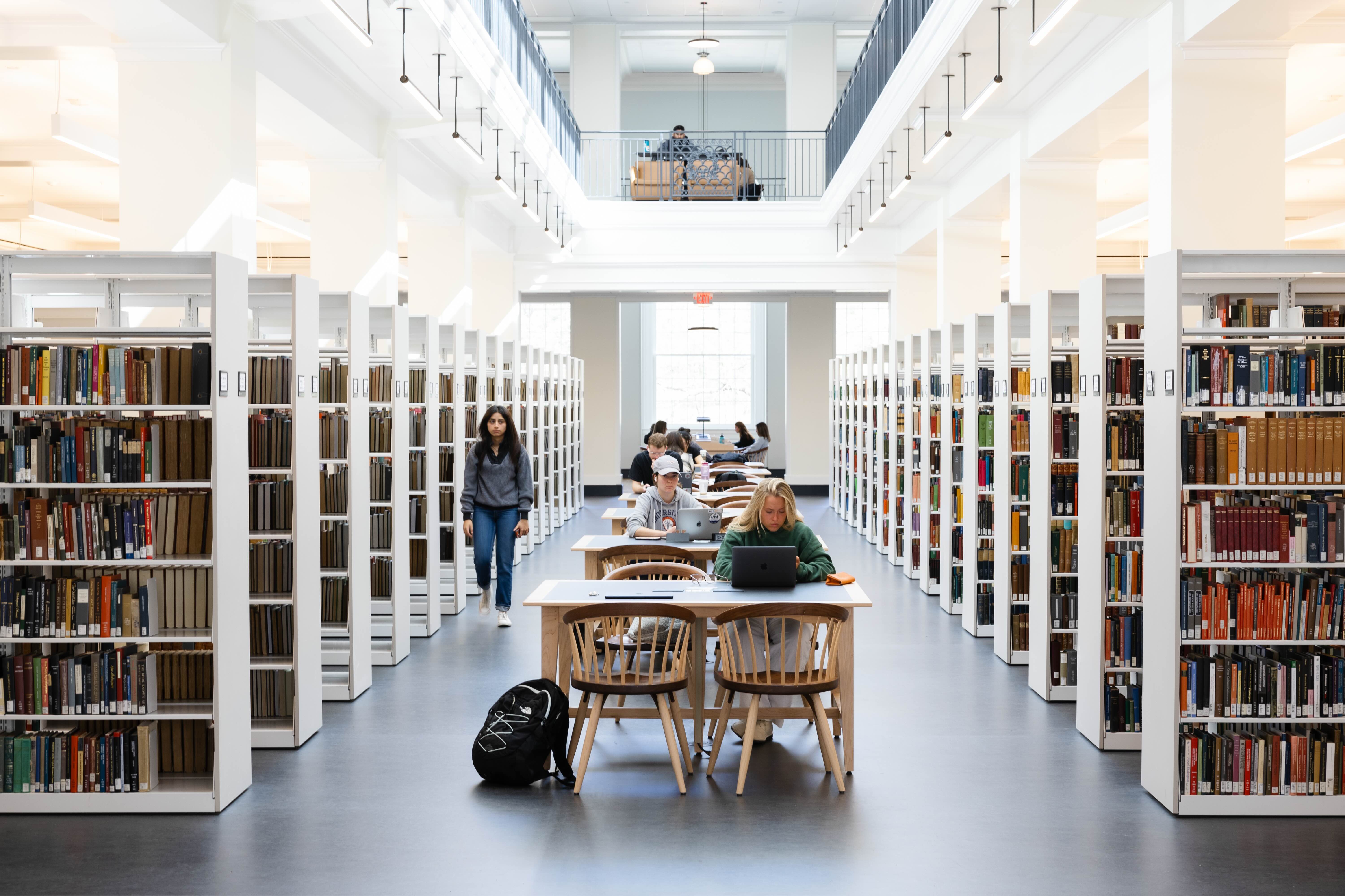 Interior of a well-lighted library with individuals reading and working at tables between rows of bookshelves.