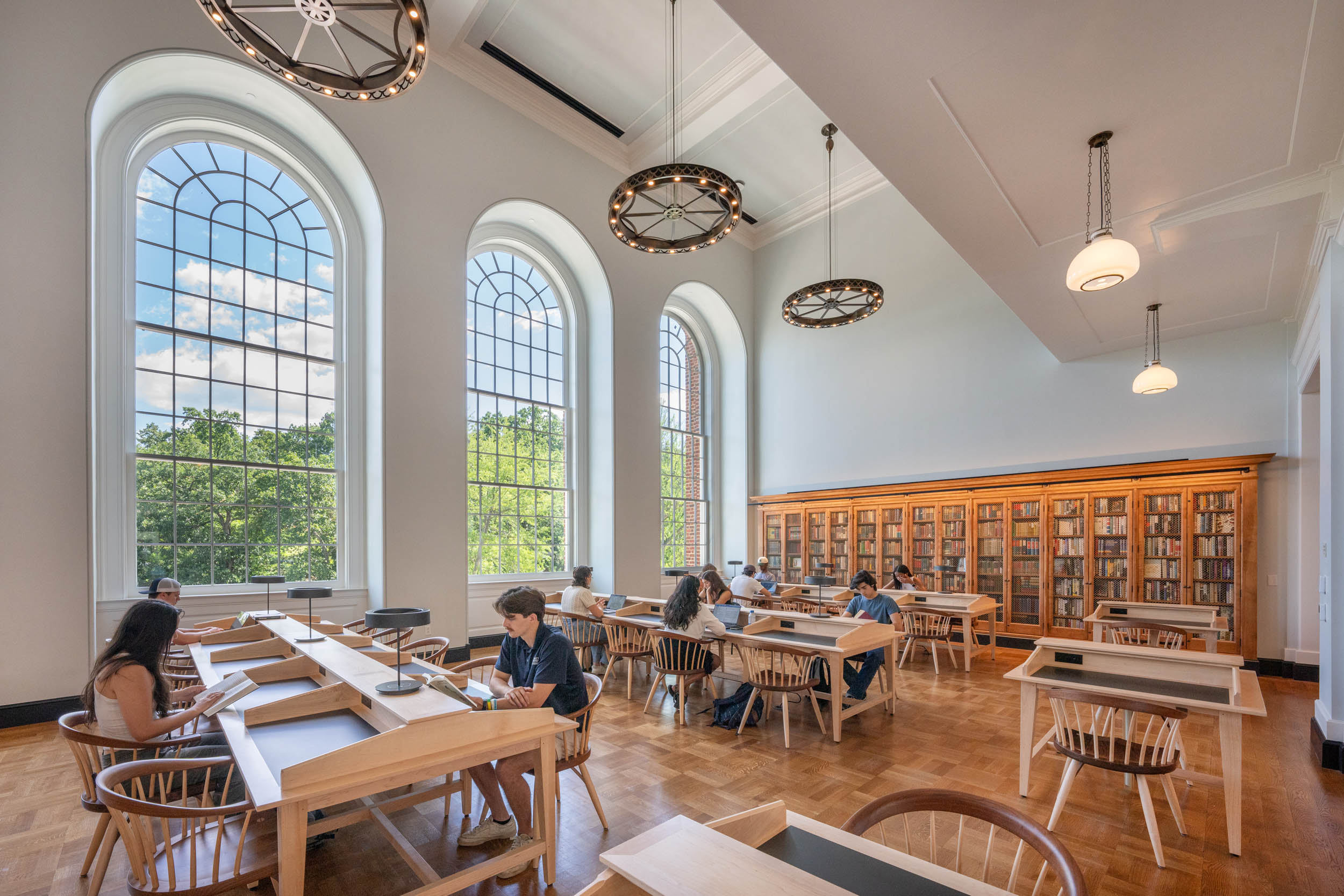 Interior of a spacious library with large arched windows, individuals seated at tables reading and using laptops, surrounded by bookshelves, with large chandeliers hanging from the ceiling.