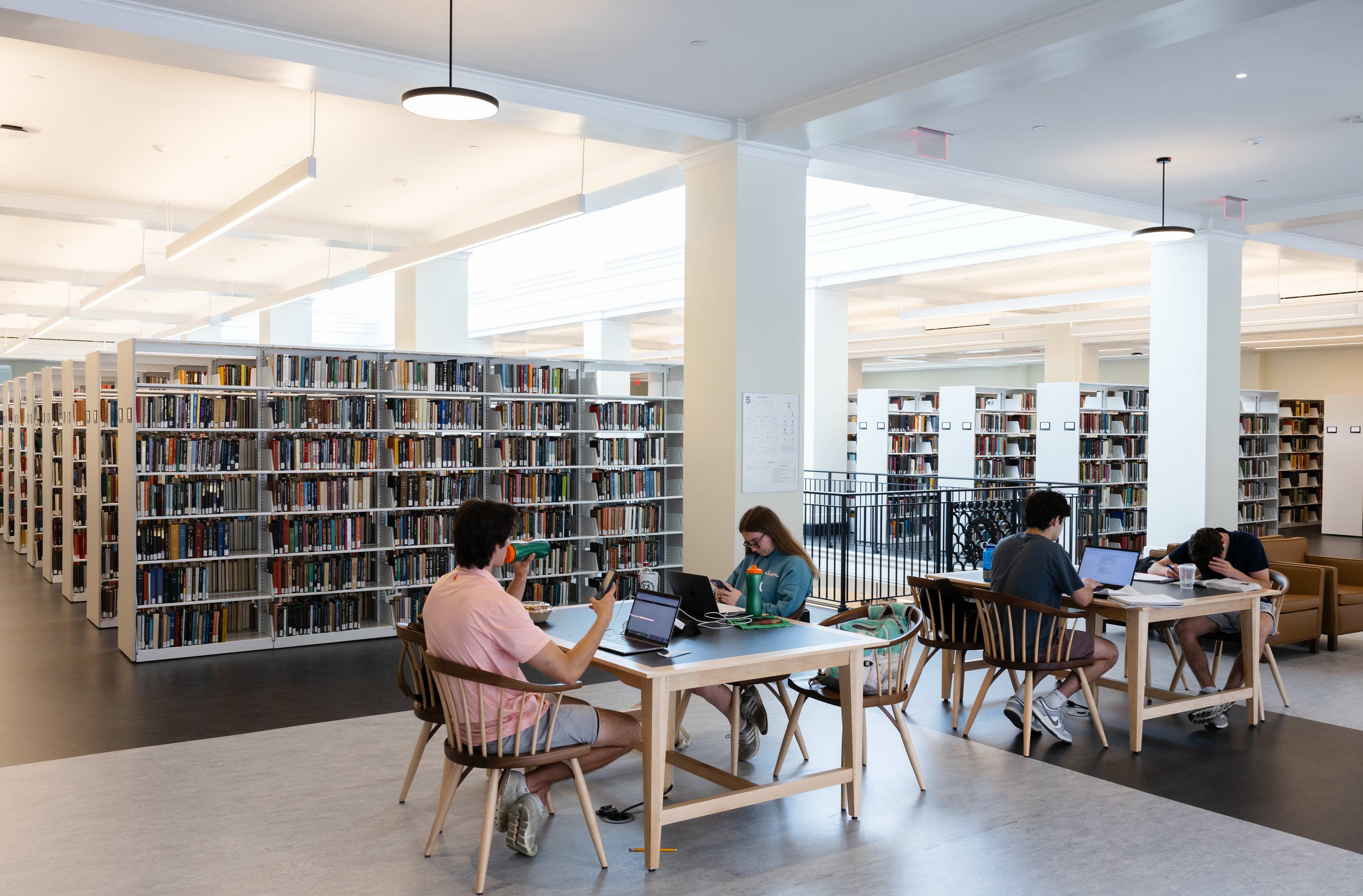 Several individuals studying and working on laptops at tables surrounded by bookshelves in a brightly lit library.