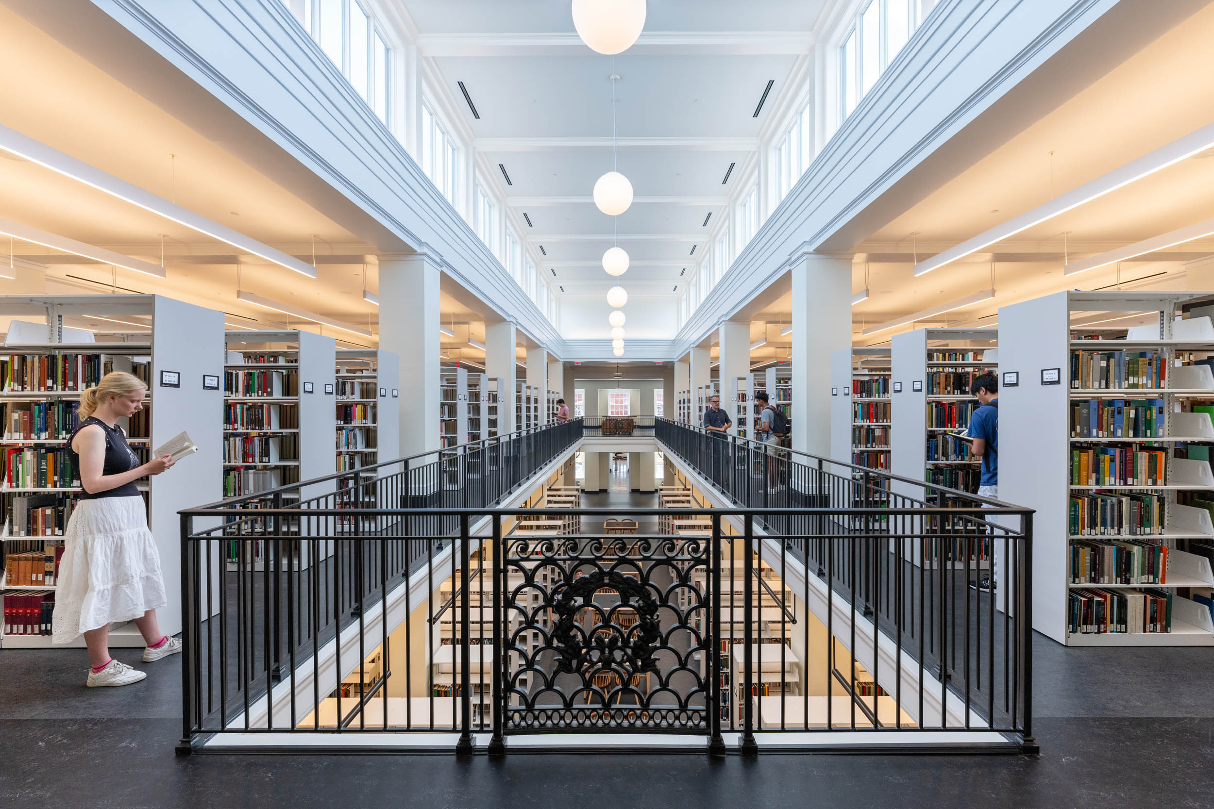 A person browsing books in the brightly lit, modern interior of a library with rows of bookshelves on either side and a central aperture surrounded by a railing.