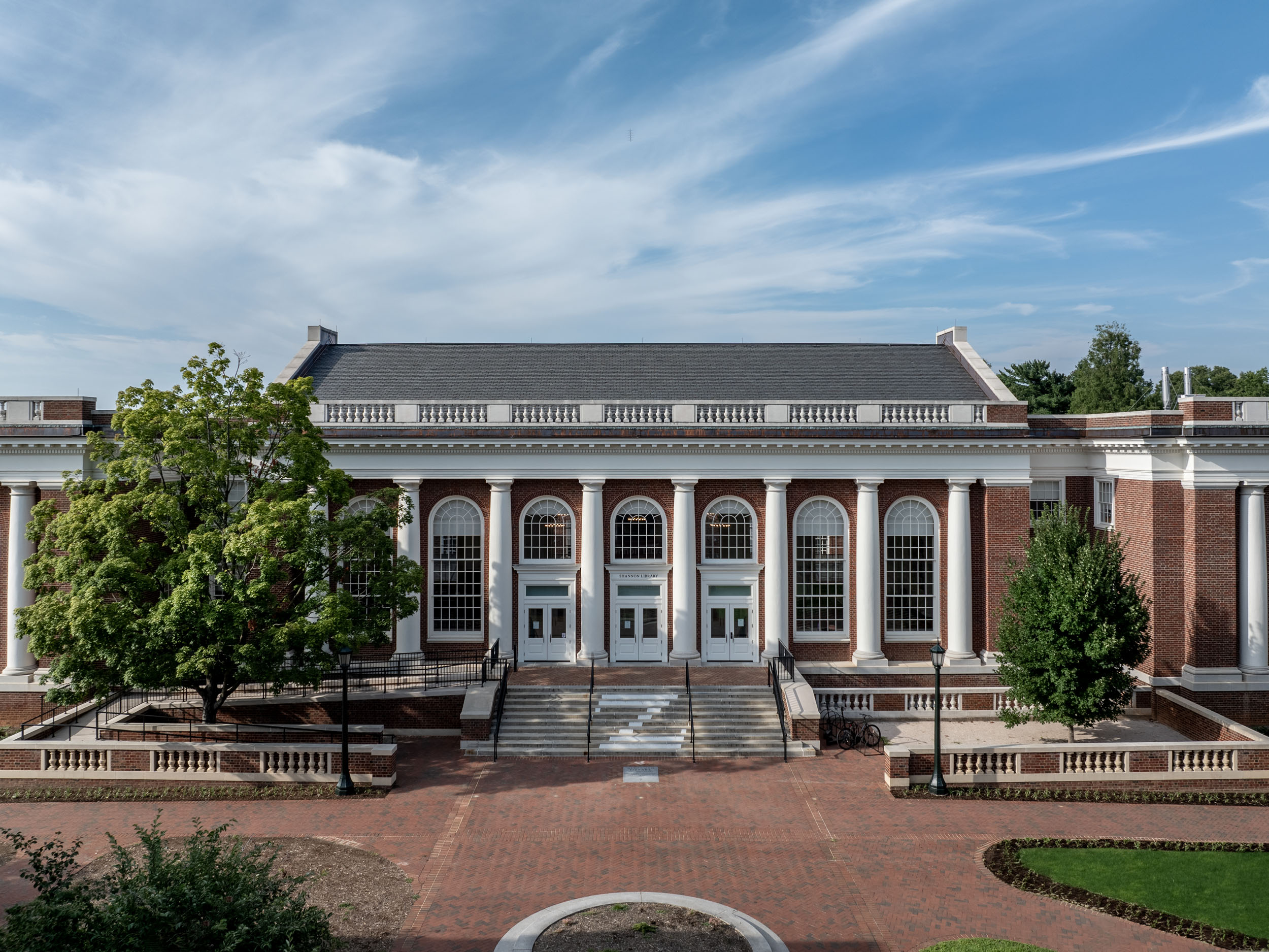 Exterior view of Shannon Library at the University of Virginia under a blue sky streaked with light clouds, featuring a brick facade with white columns and multiple arched entry doors.