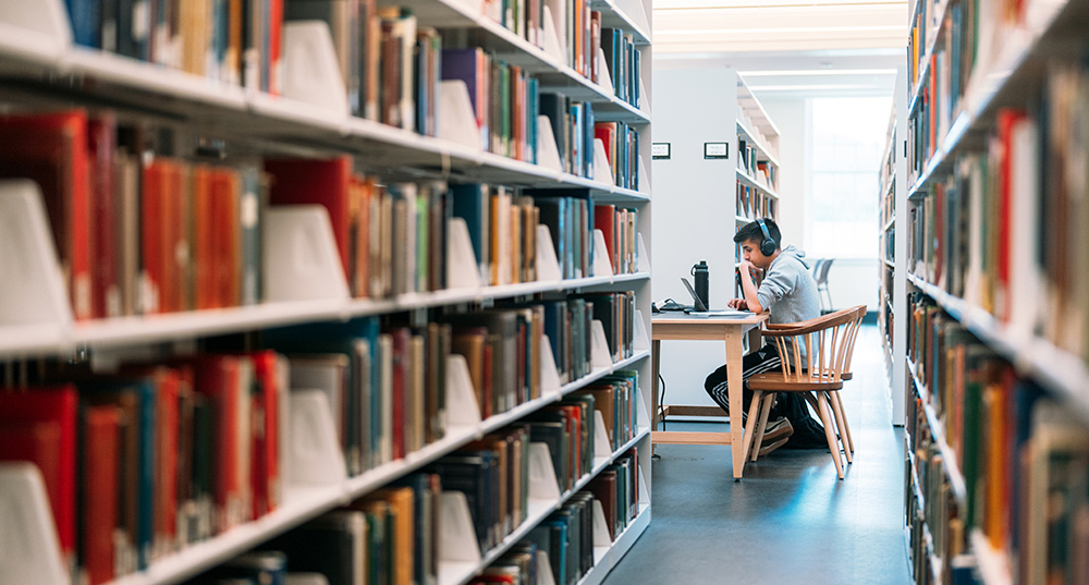 Person wearing headphones studies at a library table between tall bookshelves.