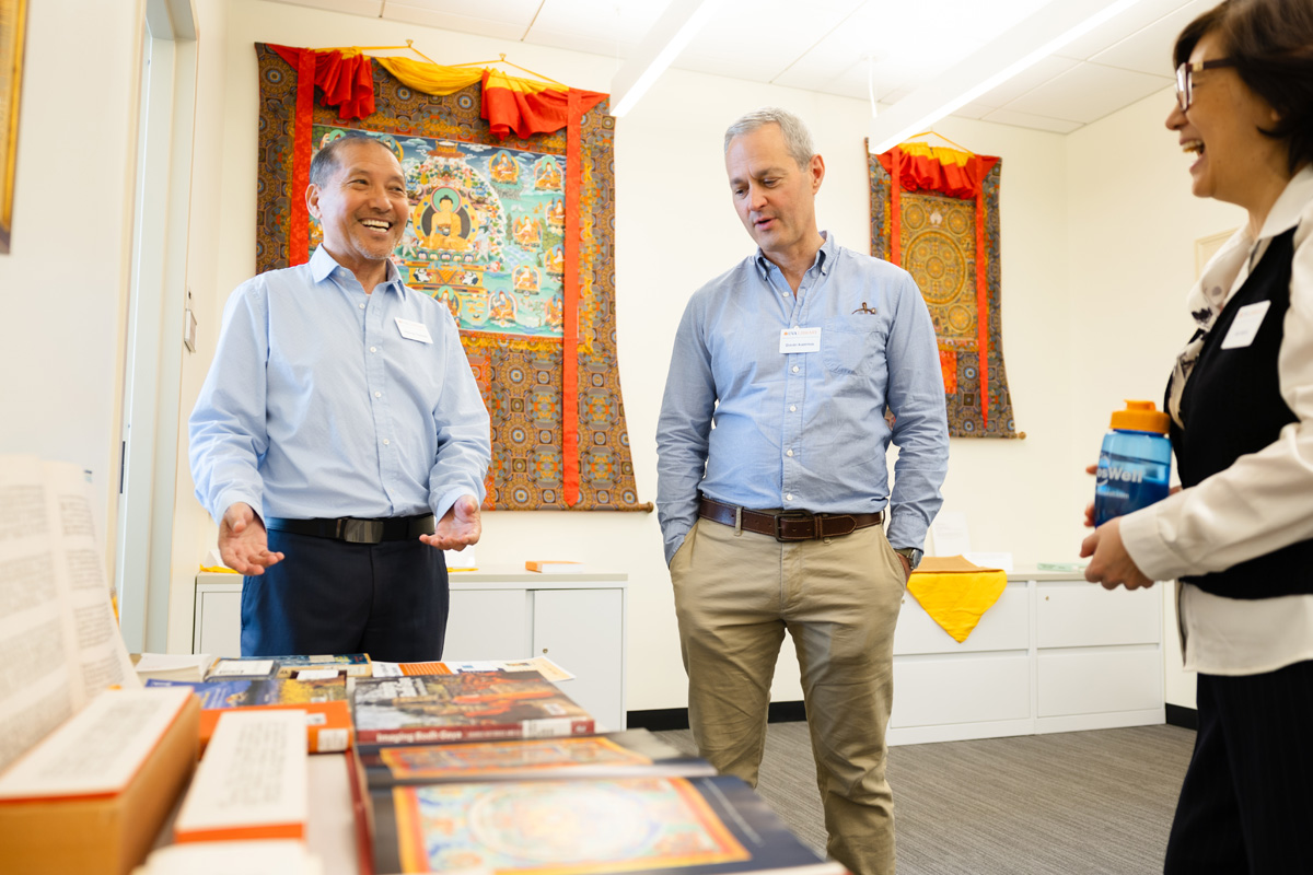 Three people are seen conversing happily in a room adorned with colorful Tibetan tapestries and a table covered with books and artifacts.