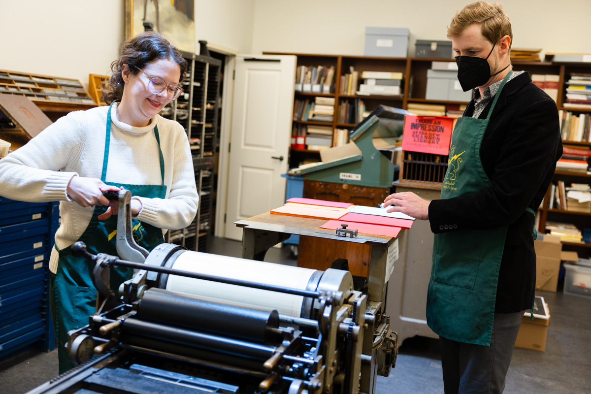 Two individuals are working with a printing press in a workshop. One person is cranking the press, while the other observes the process, both are smiling and wearing protective aprons.