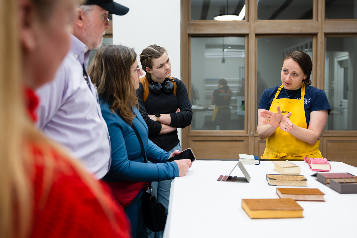 A workshop instructor in a yellow apron demonstrates a technique to a group of attentive participants around a table with various materials laid out.