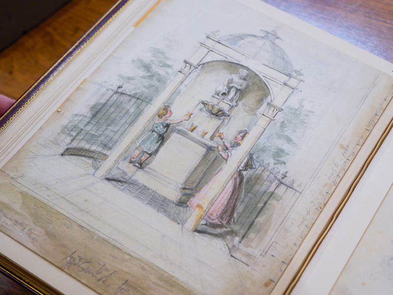 A book open to a watercolor drawing of a young woman and an adolescent filling glasses from a small fount framed by a wrought-iron fence.