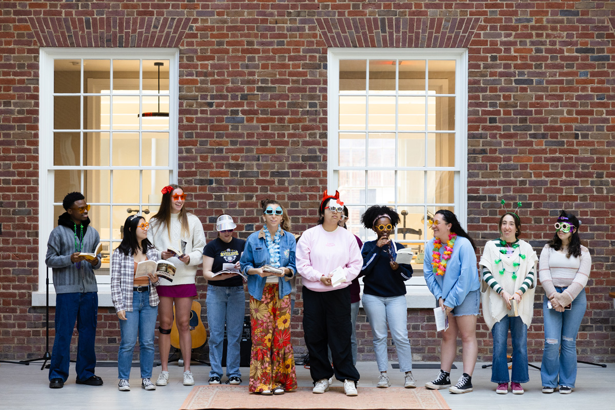 A diverse group of eleven individuals standing together in front of a brick wall, each dressed in unique and colorful casual outfits. Some are wearing hats and sunglasses, and they all appear relaxed and cheerful, enjoying an outdoor gathering.