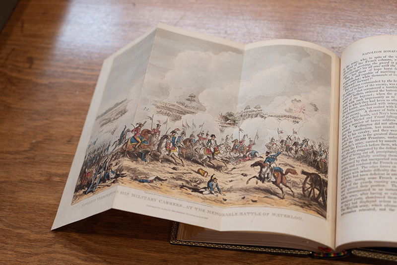 A book, open to a color page that folds out to show an illustration. The illustration is of a battle fought with horses and cannon.