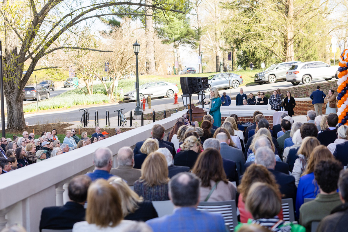 Attendees seated at an outdoor ceremony near a stage with speakers, surrounded by trees and a parking area in the background.