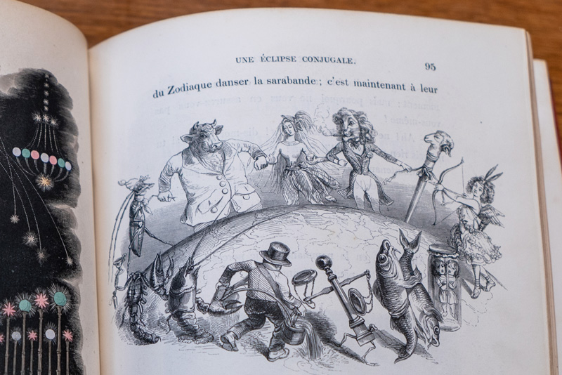 A book open to a black and white drawing of figures representing the symbols of the Zodiac. They are holding hands and apparently circling around the earth. 