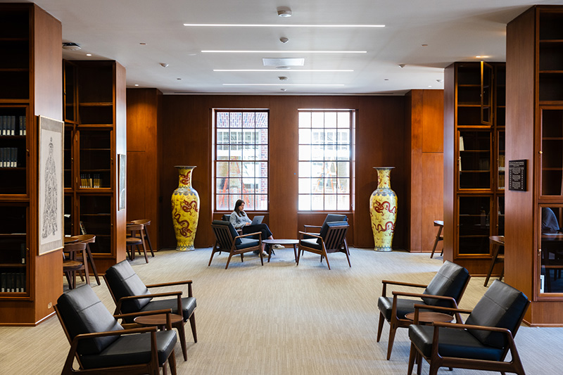 A room with shelving and mid-century modern chairs and tables. At the end of a room a person sits in a chair in front of a pair of windows between two very large yellow vases.