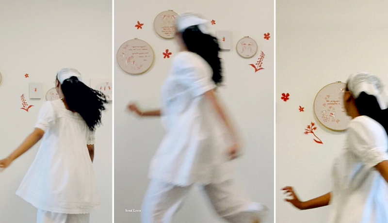 A person in white clothing is moving across a wall, which is decorated with embroidery hoops and red designs. The background has various circular embroidery pieces with different patterns. The scene has a light, airy feeling.