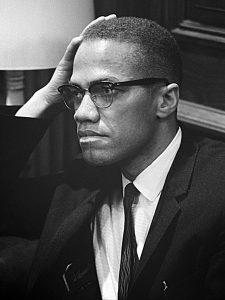 A Black man wearing glasses and a coat and tie, his hand on his head.