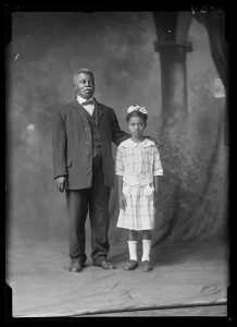 Vintage black and white photograph of an older African American man standing beside a young child, both dressed in formal clothing, in a studio setting with a painted backdrop.