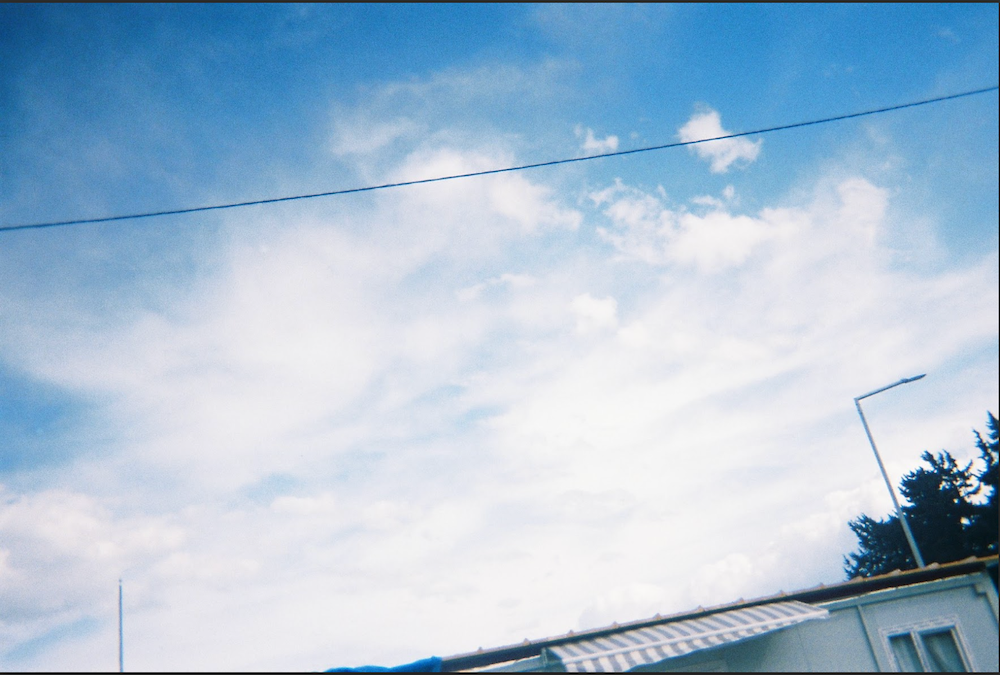 A clear blue sky with scattered white clouds. A thin overhead wire stretches across the scene. Below, part of a building with a corrugated roof is visible, alongside a streetlight and some trees.