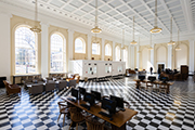A large lobby/main hall in Alderman Library. The floor is checkered, large light fixtures hang from the ceiling, sunlight streams through large windows. Tables with public computers and couches and chairs dot the room.