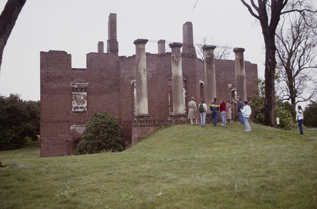 A group of people stand near the ruins of the Barboursville mansion. The structure features tall, worn brick walls and several standing columns. The grassy area surrounding the building is dotted with trees and bushes.