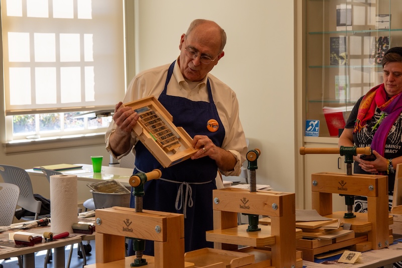 A person wearing an apron inspects a wooden tool in a workshop environment, with various woodworking tools and another person visible in the background.
