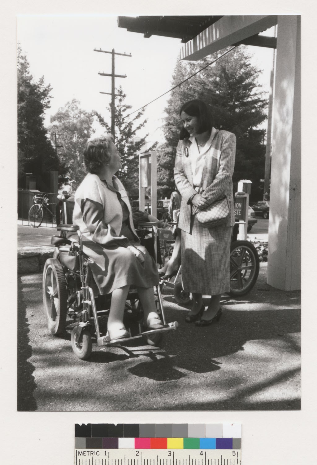 Photo from the UC Berekely Disability Rights and Independent Living Movement Collection of a person seated in a wheelchair is engaged conversation with another standing person outdoors. Both are dressed in formal attire. The scene includes trees and a bicycle in the background.