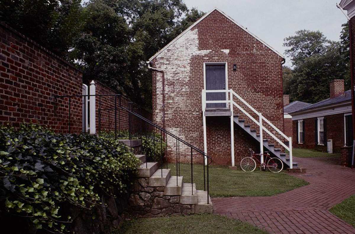 A brick building with a white staircase leading to an upper door, surrounded by red brick pathways and green foliage. A bicycle is parked near the base of the stairs. Trees are visible in the background.