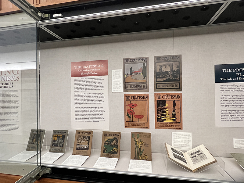 The image shows an exhibition display featuring "The Craftsman," a magazine dedicated to aesthetics and design reform. Several issues of "The Craftsman" magazine are showcased, with colorful covers that include illustrations of nature, architecture, and design elements. The display also includes an informational plaque titled "The Craftsman: Aesthetics & Reform Through Design," which provides context about the magazine's historical significance and its influence on the Arts and Crafts movement. Beneath the mounted magazine covers are open copies of various issues, each propped up on stands, allowing viewers to see more of the intricate cover art and content.