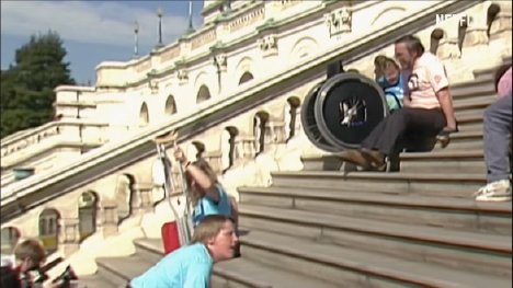 Still image from the documentary “Crip Camp” of a person in a wheelchair being assisted up a large flight of stone steps outside a building with intricate architecture. Several other individuals are also on the steps, some with crutches.