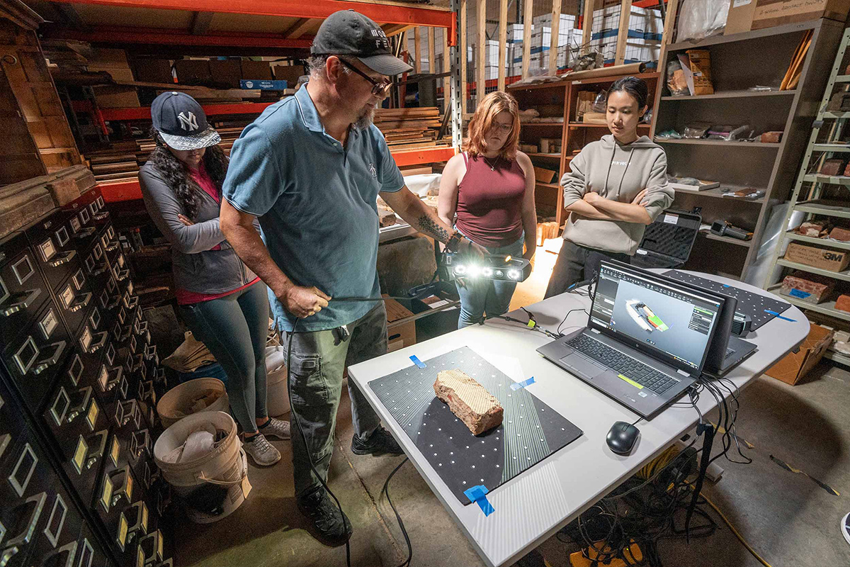 A Scholars’ Lab employee stands over a table with a brick and a laptop on it. Three students stand nearby. The group is surrounded by archival shelving.