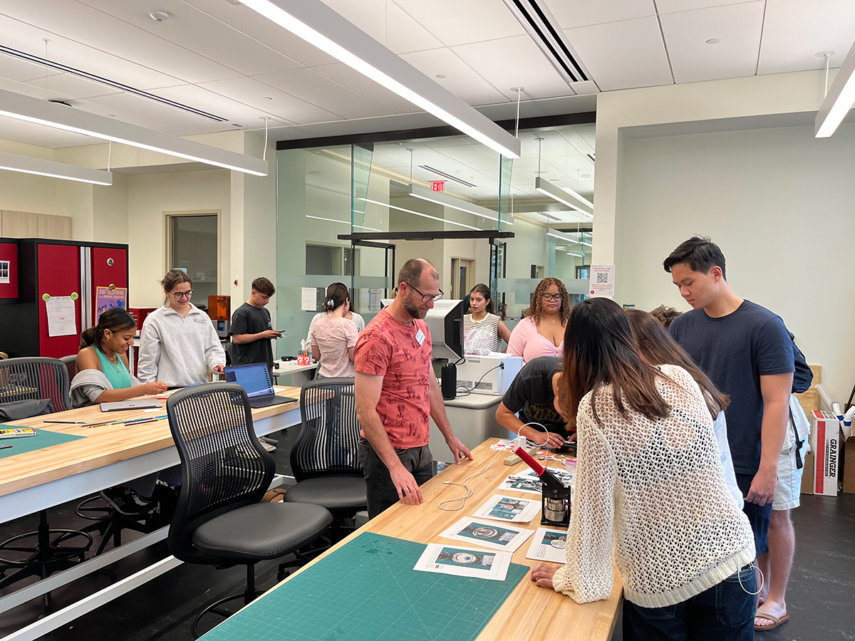 A group of community members surrounds two large work tables spread with craft and making supplies, and makerspace equipment is visible in the background.