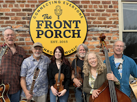 A group of six people stand in front of a brick wall with a sign that says "The Front Porch: Connecting Everyone Through Music, Est. 2013." Two hold guitars, one holds a violin, and another holds a double bass. They are smiling and outdoors.