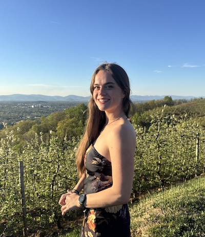 A person is standing in a vineyard with a scenic view of rolling hills and a clear blue sky in the background.