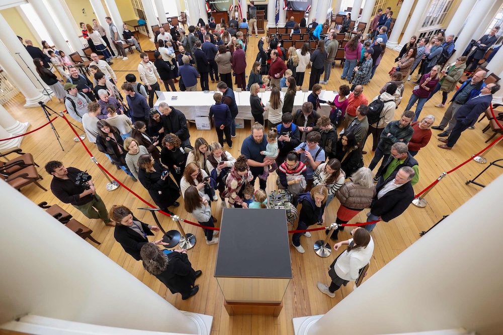 A large group of people gathers in a spacious hall with wooden flooring and tall columns. Red ropes create a barrier around an exhibit or platform in the center, with attendees observing or waiting in line. The hall features a high ceiling and natural light, creating an open ambiance.