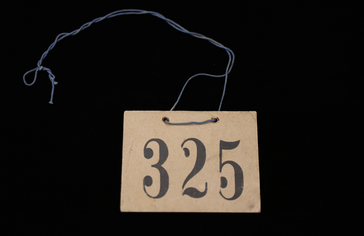 A tag with the number 325 printed in bold black font on a beige background, with a blue string attached, photographed against a black backdrop.